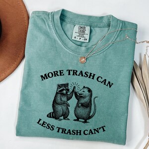 Puede incluir: Una camiseta Comfort Colors de color verde azulado con el texto "MORE TRASH CAN LESS TRASH CAN'T" y un gr&aacute;fico de un mapache y una zarig&uuml;eya chocando los cinco. Un collar dorado con un colgante est&aacute; sobre la camiseta.