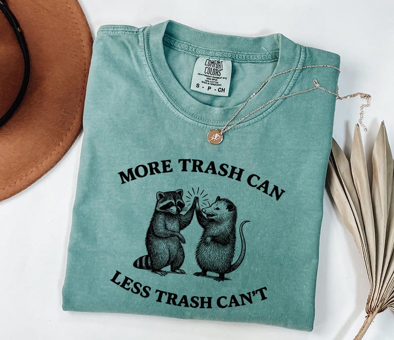 Puede incluir: Una camiseta Comfort Colors de color verde azulado con el texto "MORE TRASH CAN LESS TRASH CAN'T" y un gr&aacute;fico de un mapache y una zarig&uuml;eya chocando los cinco. Un collar dorado con un colgante est&aacute; sobre la camiseta.