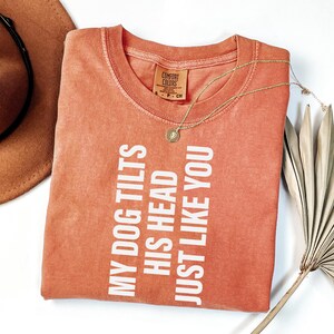 以下が含まれることがあります： 「MY DOG TILTS HIS HEAD JUST LIKE YOU」の白い文字が入った、コーラル色のTシャツ。 金色のネックレスがTシャツの上に置かれています。 茶色の帽子と乾燥したヤシの葉が背景にあります。