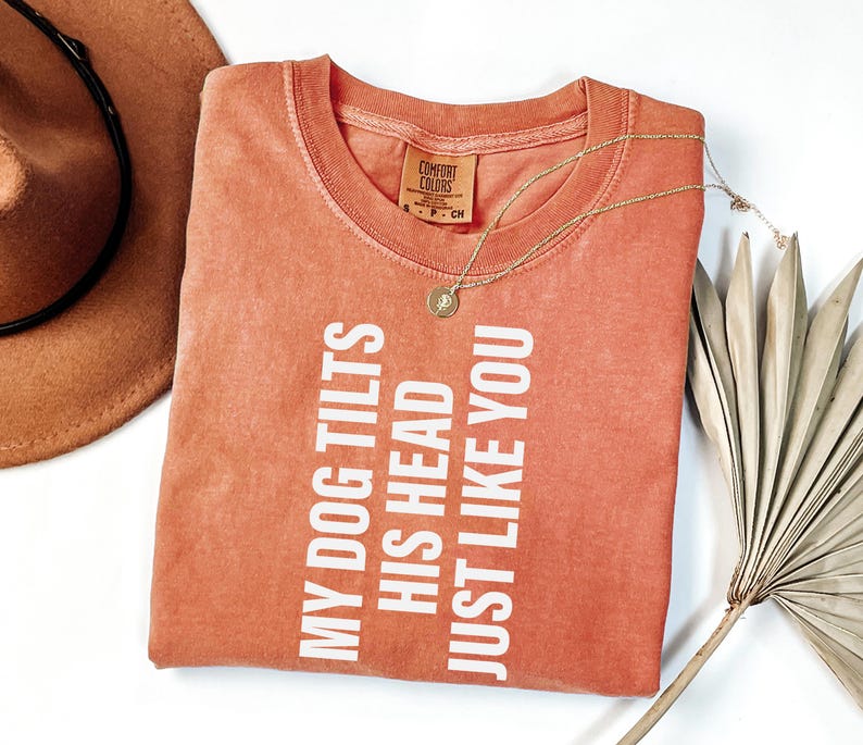 以下が含まれることがあります： 「MY DOG TILTS HIS HEAD JUST LIKE YOU」の白い文字が入った、コーラル色のTシャツ。 金色のネックレスがTシャツの上に置かれています。 茶色の帽子と乾燥したヤシの葉が背景にあります。