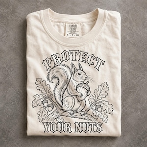 May include: A cream-colored t-shirt featuring a black line art illustration of a squirrel holding an acorn on an oak branch. The text "PROTECT YOUR NUTS" is printed in a bold, arched font above and below the squirrel graphic.