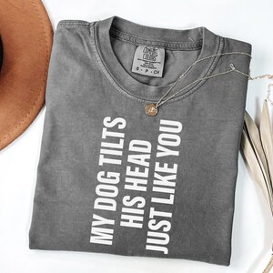 以下が含まれることがあります： 「MY DOG TILTS HIS HEAD JUST LIKE YOU」の白い文字が入ったグレーのComfort Colors Tシャツ。 金色のネックレスとペンダントがシャツの上にあります。 背景には茶色の帽子が見えます。