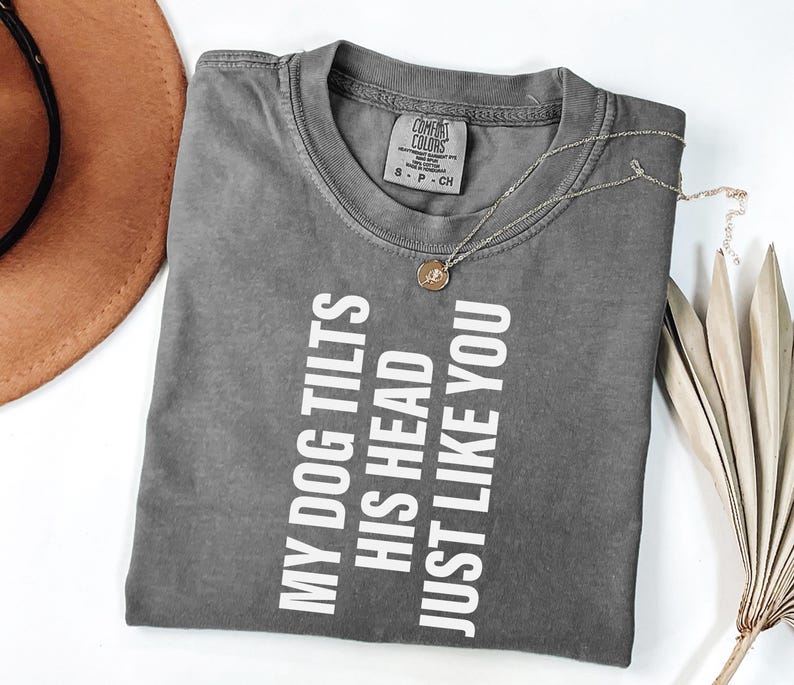 以下が含まれることがあります： 「MY DOG TILTS HIS HEAD JUST LIKE YOU」の白い文字が入ったグレーのComfort Colors Tシャツ。 金色のネックレスとペンダントがシャツの上にあります。 背景には茶色の帽子が見えます。