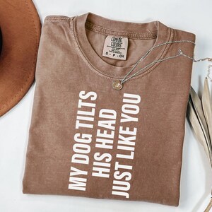 May include: A brown Comfort Colors t-shirt with the text "MY DOG TILTS HIS HEAD JUST LIKE YOU" in white. A silver necklace with a small pendant is draped across the shirt. A brown hat and dried plant are in the background.