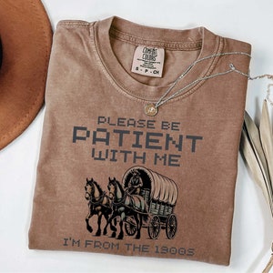 May include: A brown t-shirt with a vintage-style illustration of a horse-drawn wagon. The text "Please be patient with me I'm from the 1800s" is printed on the shirt.