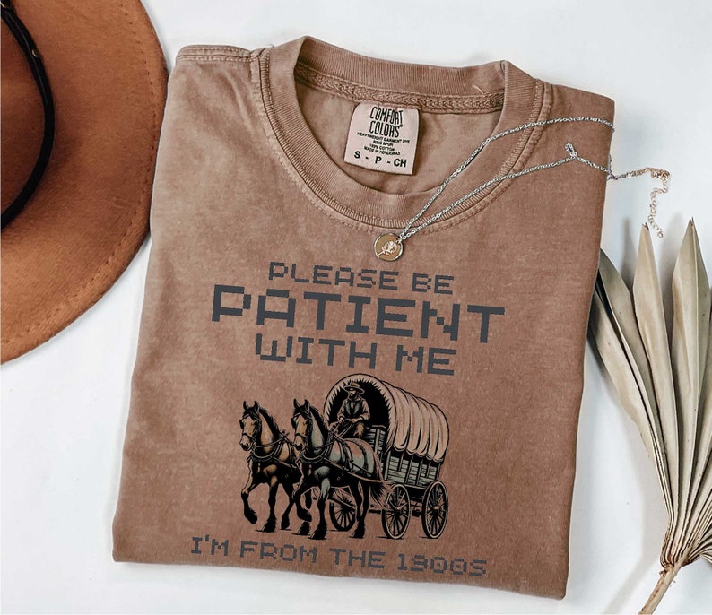 May include: A brown t-shirt with a vintage-style illustration of a horse-drawn wagon. The text "Please be patient with me I'm from the 1800s" is printed on the shirt.