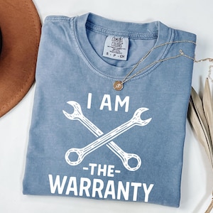 May include: A blue Comfort Colors t-shirt with the text "I AM -THE- WARRANTY" and two crossed wrenches in white. A gold necklace with a pendant is on the shirt. A brown hat and dried palm leaves are in the background.