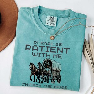 May include: A teal blue t-shirt with a vintage-style graphic of a horse-drawn wagon. The text "PLEASE BE PATIENT WITH ME I'M FROM THE 1900S" is printed in black on the shirt.