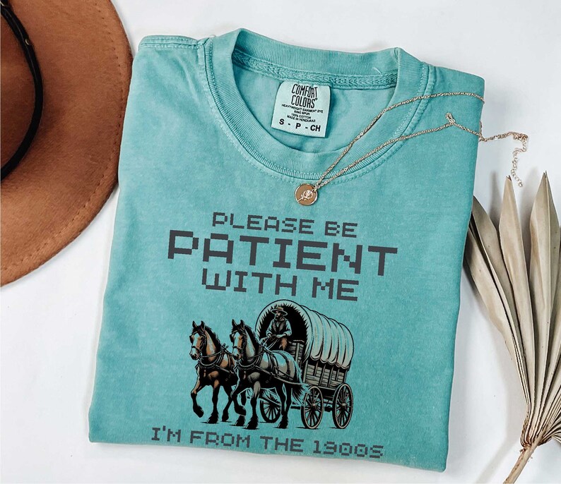 May include: A teal blue t-shirt with a vintage-style graphic of a horse-drawn wagon. The text "PLEASE BE PATIENT WITH ME I'M FROM THE 1900S" is printed in black on the shirt.