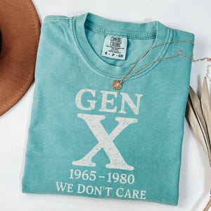 May include: A turquoise t-shirt with the text "GEN X 1965-1980 WE DON'T CARE" in white. A gold necklace with a pendant is draped over the shirt. A brown hat is visible in the background.