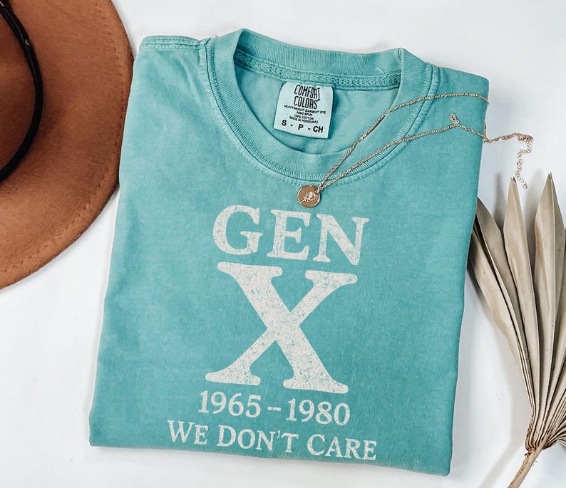 May include: A turquoise t-shirt with the text "GEN X 1965-1980 WE DON'T CARE" in white. A gold necklace with a pendant is draped over the shirt. A brown hat is visible in the background.