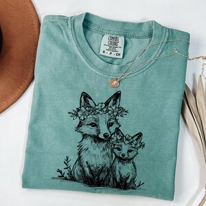 May include: A seafoam green Comfort Colors t-shirt featuring a black ink illustration of two foxes wearing floral crowns. A delicate silver necklace with a small pendant rests on the shirt. The shirt is folded, with a brown hat and dried palm leaves in the background.