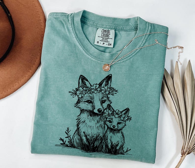 May include: A seafoam green Comfort Colors t-shirt featuring a black ink illustration of two foxes wearing floral crowns. A delicate silver necklace with a small pendant rests on the shirt. The shirt is folded, with a brown hat and dried palm leaves in the background.