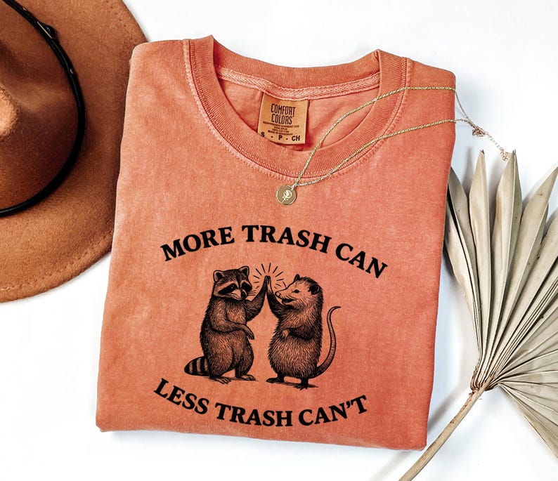 Puede incluir: Camiseta de color coral con el texto "MORE TRASH CAN LESS TRASH CAN'T" y un gr&aacute;fico de un mapache y una zarig&uuml;eya chocando los cinco. Un collar dorado y un sombrero marr&oacute;n tambi&eacute;n est&aacute;n en la imagen.