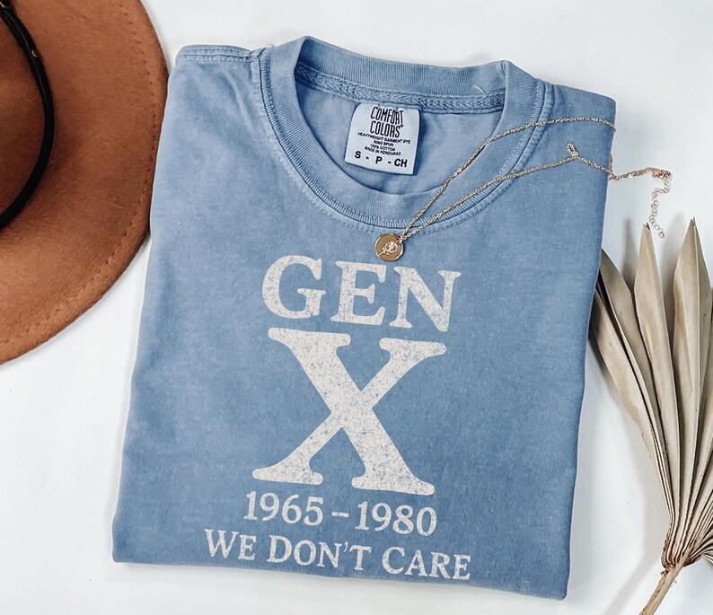 May include: A light blue t-shirt with the text "GEN X 1965-1980 WE DON'T CARE" in white. The shirt is folded and has a small tag on the collar. A gold necklace with a pendant is draped across the shirt. A brown hat and dried palm leaves are in the background.