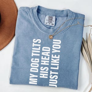 以下が含まれることがあります： 「MY DOG TILTS HIS HEAD JUST LIKE YOU」の白い文字が入った青いTシャツ。ペンダント付きのゴールドネックレスがシャツの上にあります。シャツは白い表面に折りたたまれています。