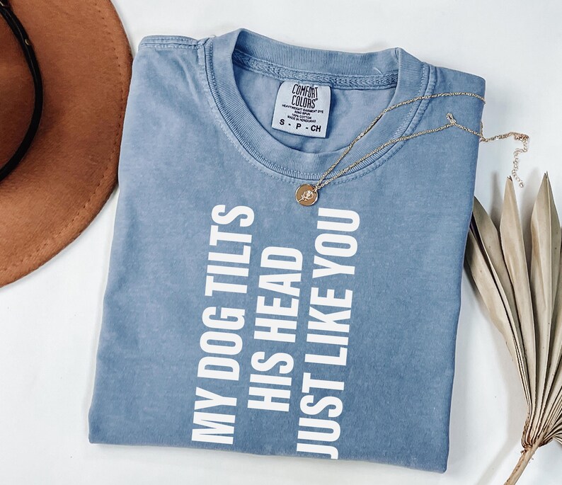 以下が含まれることがあります： 「MY DOG TILTS HIS HEAD JUST LIKE YOU」の白い文字が入った青いTシャツ。ペンダント付きのゴールドネックレスがシャツの上にあります。シャツは白い表面に折りたたまれています。