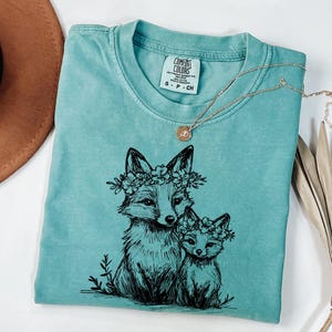 May include: A turquoise Comfort Colors t-shirt featuring a black ink illustration of two foxes wearing floral crowns. A gold necklace with a pendant rests on the shirt. The shirt is folded, with a brown hat visible in the background.
