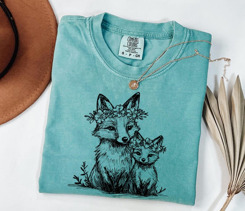 May include: A turquoise Comfort Colors t-shirt featuring a black ink illustration of two foxes wearing floral crowns. A gold necklace with a pendant rests on the shirt. The shirt is folded, with a brown hat visible in the background.