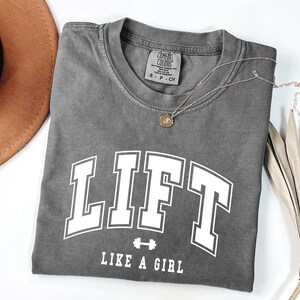 Pode incluir: Uma camiseta cinza com a palavra "LIFT" em letras brancas grandes e a frase "LIKE A GIRL" abaixo. Um pequeno gr&aacute;fico de barra est&aacute; entre as palavras. Um colar de ouro com um pingente est&aacute; na camisa. Um chap&eacute;u marrom est&aacute; no fundo.
