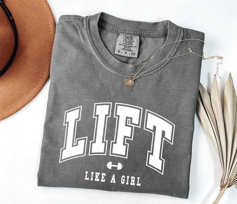 Pode incluir: Uma camiseta cinza com a palavra "LIFT" em letras brancas grandes e a frase "LIKE A GIRL" abaixo. Um pequeno gr&aacute;fico de barra est&aacute; entre as palavras. Um colar de ouro com um pingente est&aacute; na camisa. Um chap&eacute;u marrom est&aacute; no fundo.