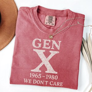 May include: A red t-shirt with the text "GEN X 1965-1980 WE DON'T CARE" in white block letters. The shirt has a crew neck and short sleeves. A gold necklace with a pendant is draped across the shirt. A brown hat and dried plant are in the background.