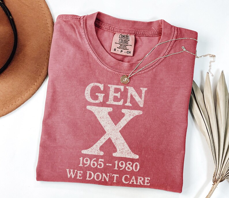 May include: A red t-shirt with the text "GEN X 1965-1980 WE DON'T CARE" in white block letters. The shirt has a crew neck and short sleeves. A gold necklace with a pendant is draped across the shirt. A brown hat and dried plant are in the background.