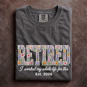May include: A charcoal gray crew neck t-shirt featuring the word RETIRED in large block letters filled with a colorful floral pattern. Below the text, the shirt displays the phrase I worked my whole life for this and Est. 2026 in white.