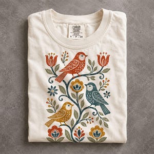 May include: A cream-colored short-sleeve t-shirt with a Comfort Colors label at the neck. The front features a graphic print of three birds in red, blue, and yellow, perched among stylized dark blue vines, leaves, and orange and blue flowers.
