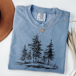 May include: A light blue Comfort Colors t-shirt featuring a black graphic of pine trees. The shirt has a crew neck and short sleeves. A gold necklace with a pendant is draped across the shirt. A brown hat and dried plant are in the background.