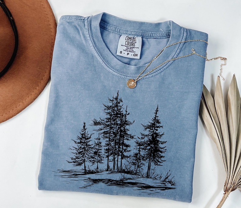 May include: A light blue Comfort Colors t-shirt featuring a black graphic of pine trees. The shirt has a crew neck and short sleeves. A gold necklace with a pendant is draped across the shirt. A brown hat and dried plant are in the background.