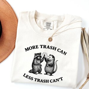 Puede incluir: Camiseta color crema con el texto "MORE TRASH CAN LESS TRASH CAN'T" y un gr&aacute;fico de un mapache y una zarig&uuml;eya chocando los cinco. La camiseta tiene cuello redondo y mangas cortas. En la imagen tambi&eacute;n hay un collar dorado y un sombrero marr&oacute;n.