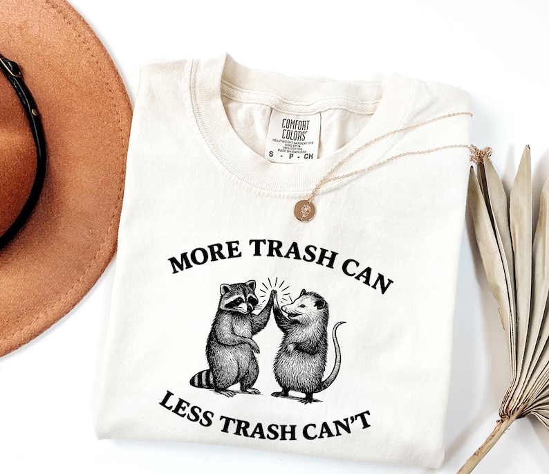 Puede incluir: Camiseta color crema con el texto "MORE TRASH CAN LESS TRASH CAN'T" y un gr&aacute;fico de un mapache y una zarig&uuml;eya chocando los cinco. La camiseta tiene cuello redondo y mangas cortas. En la imagen tambi&eacute;n hay un collar dorado y un sombrero marr&oacute;n.