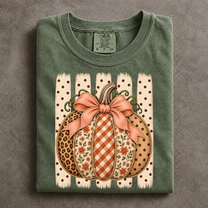 May include: An olive green t-shirt featuring a Comfort Colors label at the neck. The graphic displays a pumpkin with segments of leopard print, orange gingham, and floral patterns, topped with a large peach-colored bow over vertical stripes.