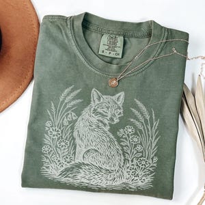 A flat-lay photograph of a sage green Comfort Colors t-shirt featuring a white line-art illustration of a fox sitting in a meadow. The fox is detailed with textured fur and is surrounded by tall grass, wildflowers, and wheat stalks. The shirt is styled on a white surface with a brown wide-brimmed hat, a gold pendant necklace, and dried palm leaves.