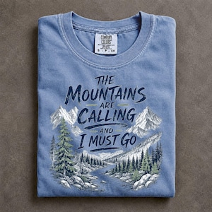 May include: A blue short-sleeved t-shirt featuring a graphic of a mountain landscape with a river and pine trees. The text reads, "THE MOUNTAINS ARE CALLING AND I MUST GO." The shirt has a crew neckline and a Comfort Colors brand label at the neck.