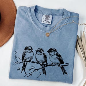 May include: A light blue Comfort Colors t-shirt featuring a black illustration of three birds perched on a branch. A gold necklace with a small pendant rests on the shirt. A brown hat is visible in the upper left corner.
