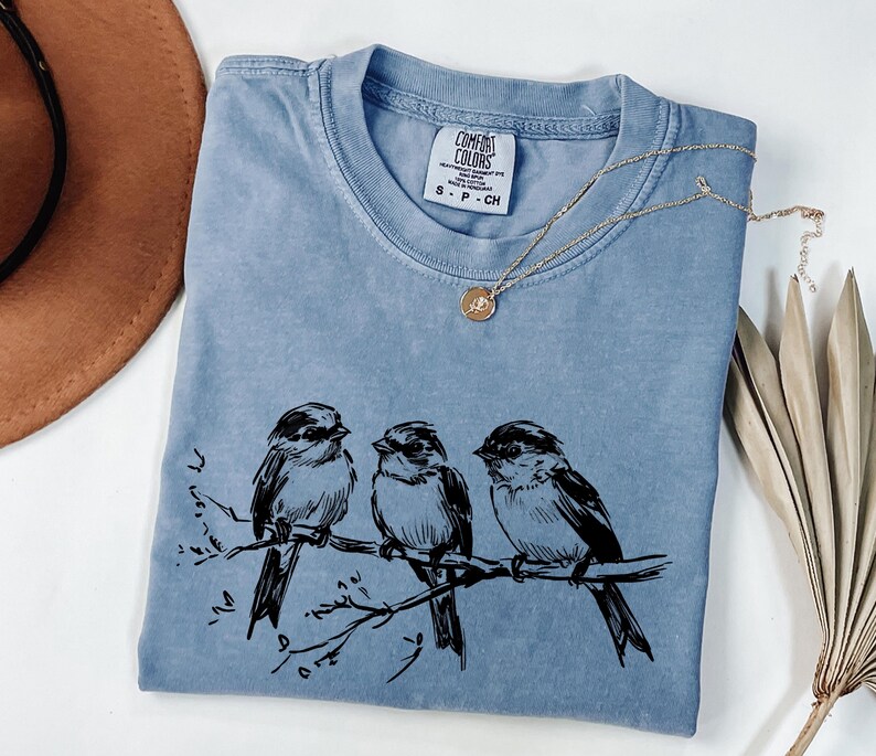 May include: A light blue Comfort Colors t-shirt featuring a black illustration of three birds perched on a branch. A gold necklace with a small pendant rests on the shirt. A brown hat is visible in the upper left corner.