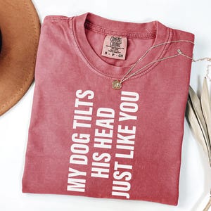 以下が含まれることがあります： 「MY DOG TILTS HIS HEAD JUST LIKE YOU」の白文字がプリントされた赤いTシャツ。クルーネックと半袖です。金色のペンダント付きネックレスがTシャツの上にあります。茶色の帽子と乾燥した葉が背景にあります。