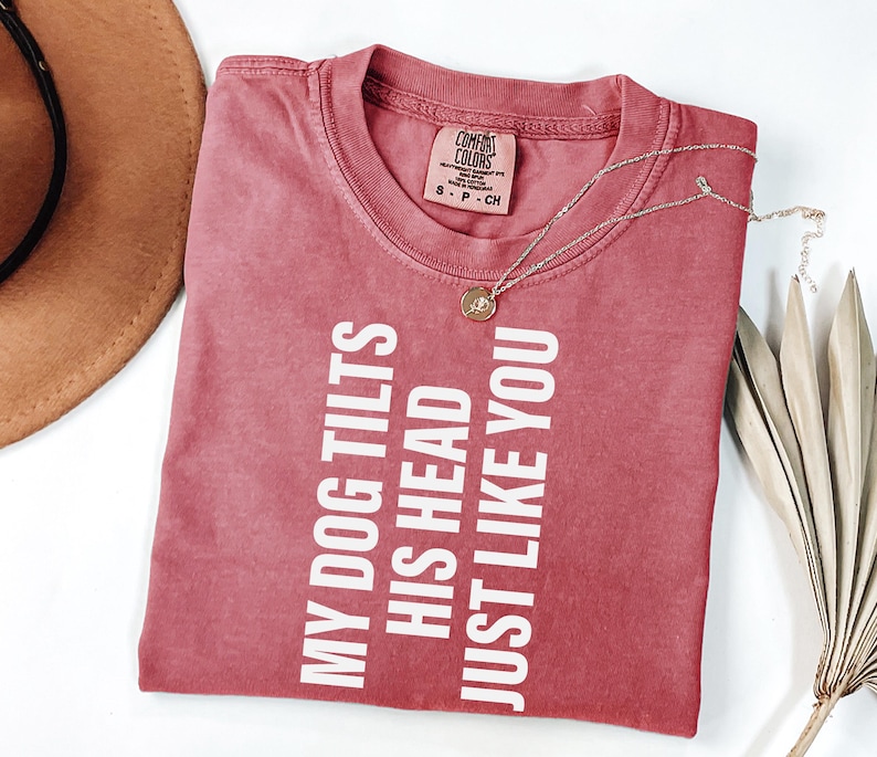 以下が含まれることがあります： 「MY DOG TILTS HIS HEAD JUST LIKE YOU」の白文字がプリントされた赤いTシャツ。クルーネックと半袖です。金色のペンダント付きネックレスがTシャツの上にあります。茶色の帽子と乾燥した葉が背景にあります。