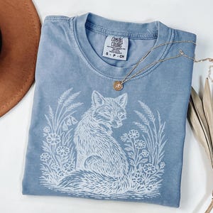 May include: A blue Comfort Colors t-shirt with a white fox graphic surrounded by flowers and foliage. The shirt has a crew neck and short sleeves. A gold necklace with a pendant is draped across the shirt.