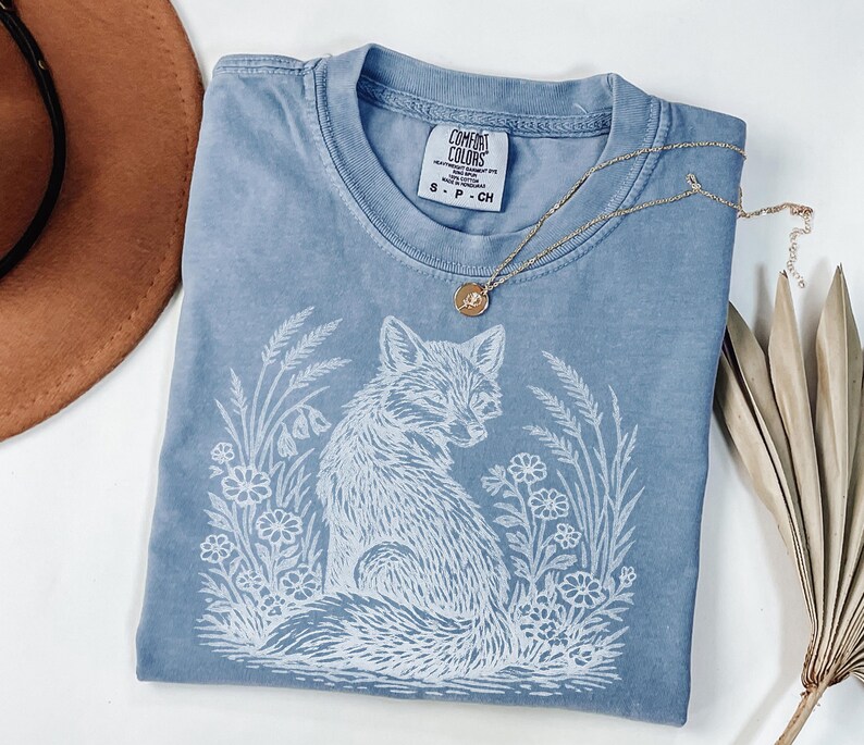 May include: A blue Comfort Colors t-shirt with a white fox graphic surrounded by flowers and foliage. The shirt has a crew neck and short sleeves. A gold necklace with a pendant is draped across the shirt.