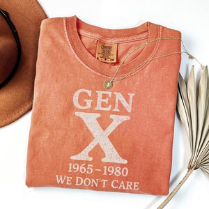 May include: Coral-colored t-shirt with the text "GEN X 1965-1980 WE DON'T CARE" in white. A gold necklace with a pendant is draped across the shirt. A brown hat and a dried palm leaf are also in the image.