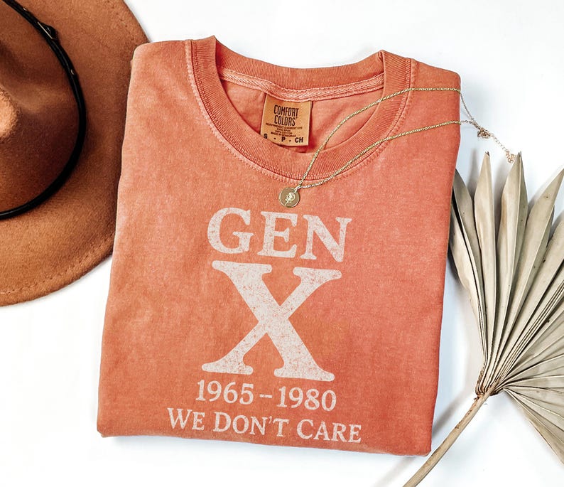 May include: Coral-colored t-shirt with the text "GEN X 1965-1980 WE DON'T CARE" in white. A gold necklace with a pendant is draped across the shirt. A brown hat and a dried palm leaf are also in the image.