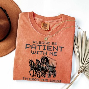 May include: A burnt orange t-shirt with the text "Please be patient with me I'm from the 1900s" in black pixelated font. A black and white illustration of a horse-drawn wagon is centered on the shirt.