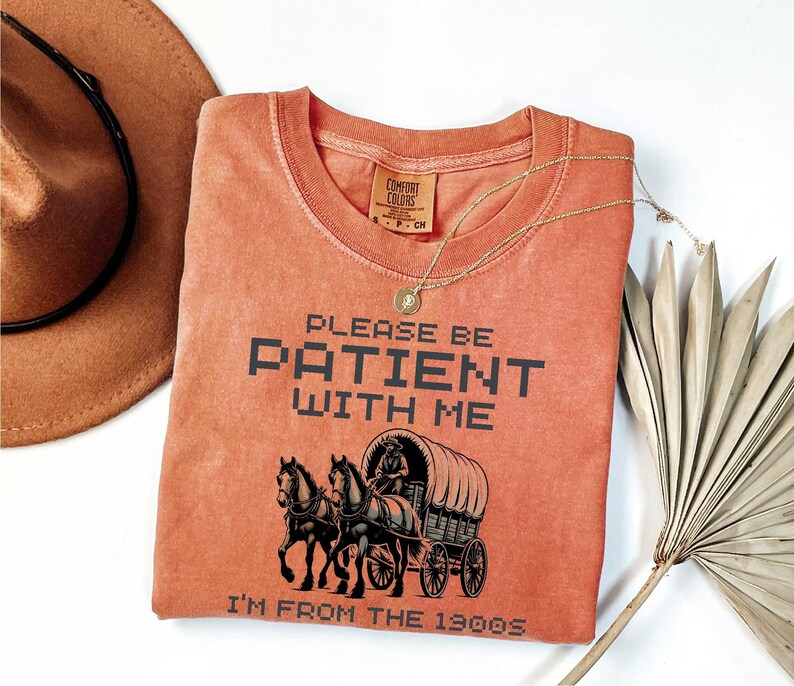 May include: A burnt orange t-shirt with the text "Please be patient with me I'm from the 1900s" in black pixelated font. A black and white illustration of a horse-drawn wagon is centered on the shirt.