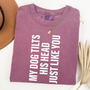以下が含まれることがあります： 「MY DOG TILTS HIS HEAD JUST LIKE YOU」の白文字が入った、モーヴ色のTシャツ。金のネックレスと茶色の帽子も見える。シャツは白い表面にきれいに折りたたまれている。