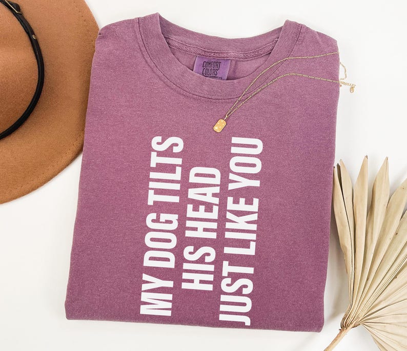 以下が含まれることがあります： 「MY DOG TILTS HIS HEAD JUST LIKE YOU」の白文字が入った、モーヴ色のTシャツ。金のネックレスと茶色の帽子も見える。シャツは白い表面にきれいに折りたたまれている。
