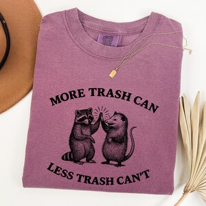 Puede incluir: Camiseta Comfort Colors malva con cuello redondo. La camiseta presenta un gr&aacute;fico de un mapache y una zarig&uuml;eya d&aacute;ndose la mano, con el texto "MORE TRASH CAN LESS TRASH CAN'T" en negro.