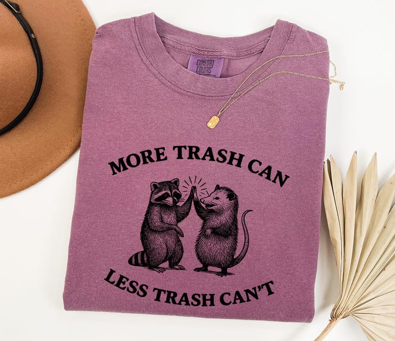 Puede incluir: Camiseta Comfort Colors malva con cuello redondo. La camiseta presenta un gr&aacute;fico de un mapache y una zarig&uuml;eya d&aacute;ndose la mano, con el texto "MORE TRASH CAN LESS TRASH CAN'T" en negro.